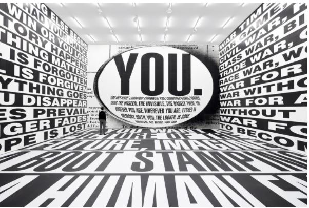 Picture of a large mirror with the word YOU in all caps in the middle at the end of a long room, words in black and white encircle the walls and floor